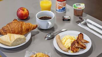 Una tavola imbandita per la colazione, con croissant, una porzione di bacon e uova, un bicchiere di succo d'arancia, una lattina di Mambo, un piccolo pacchetto di burro e una tazza di caffè.