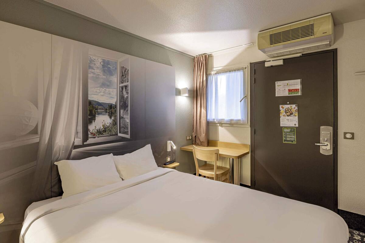 A hotel room with a bed, a desk, a chair, a window with curtains, and an air conditioning unit.