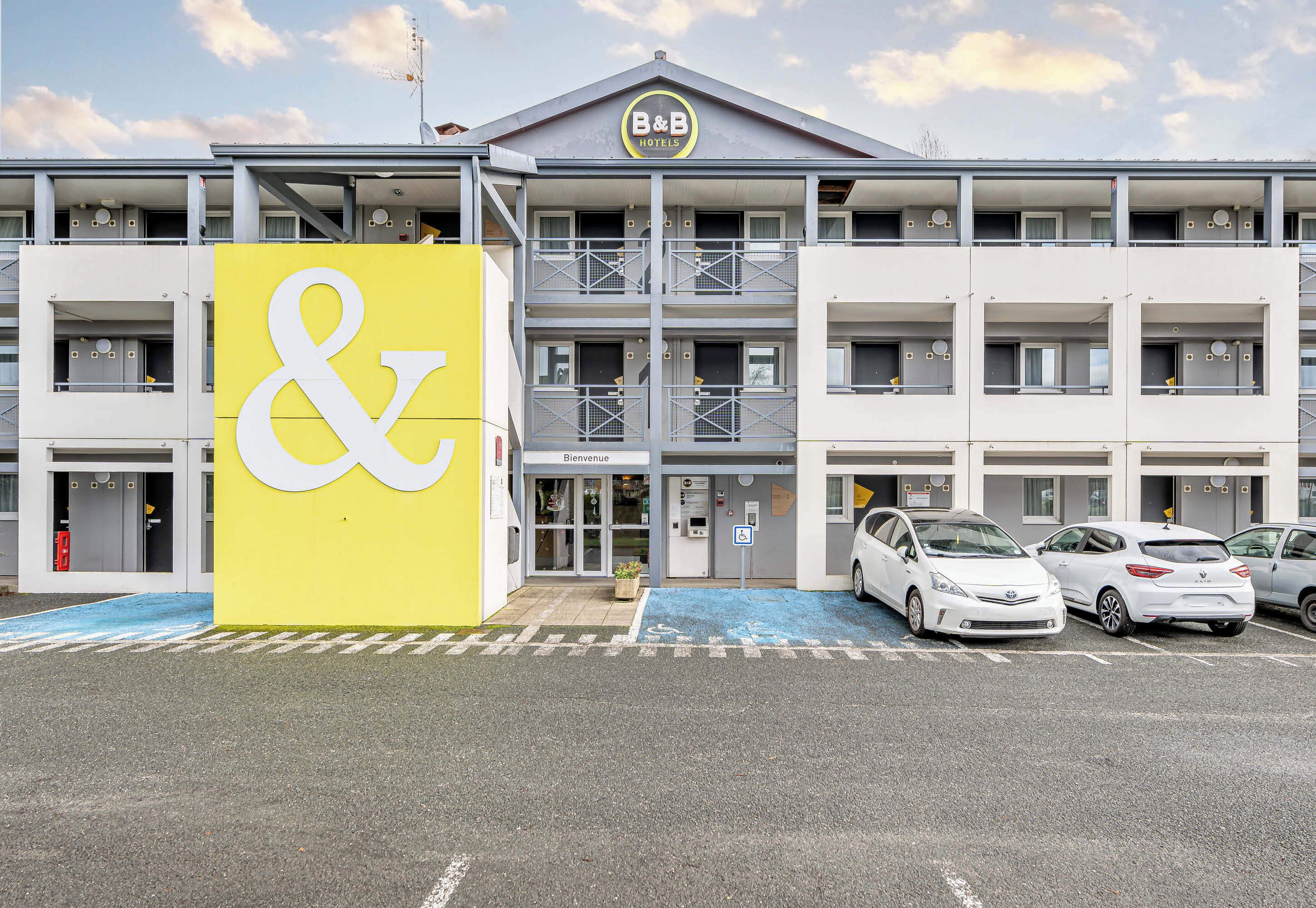 A hotel with a prominent yellow sign featuring a white ampersand symbol, multiple floors, balconies, and a parking area with several cars.