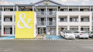 Un hôtel reconnaissable à son grand panneau jaune orné d’un symbole « et » blanc, avec de nombreux étages, des balcons et un parking où sont stationnées plusieurs voitures.