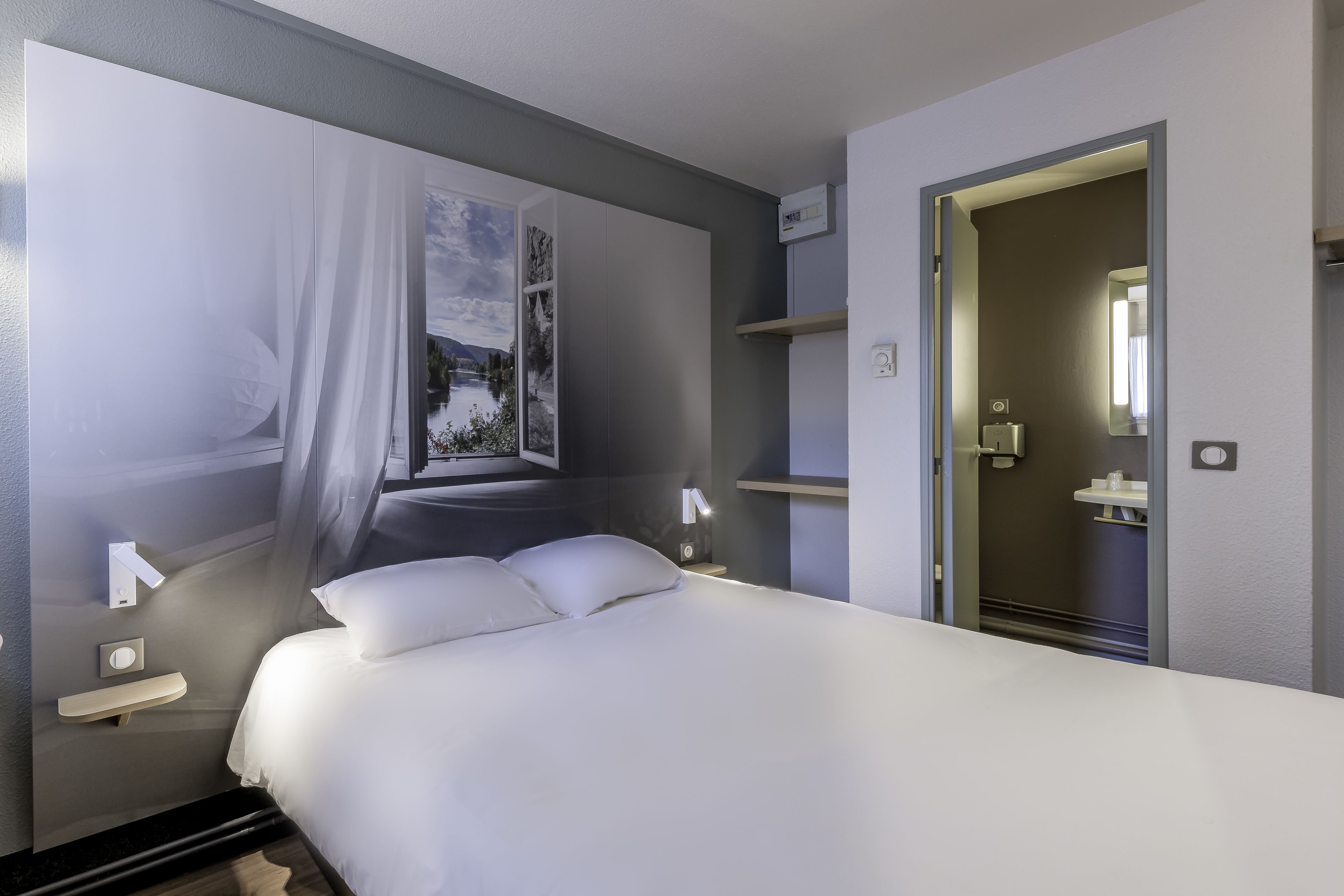 A modern hotel room with a large bed, a window with a view of a landscape, a bedside table with a lamp, and a door leading to a bathroom.