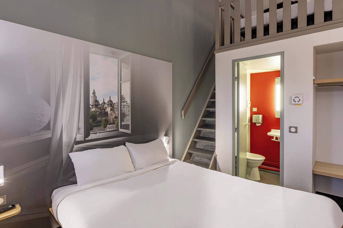 A hotel room with a bed, a nightstand, a window with sheer curtains, a staircase leading to an upper level, and a red-walled bathroom.