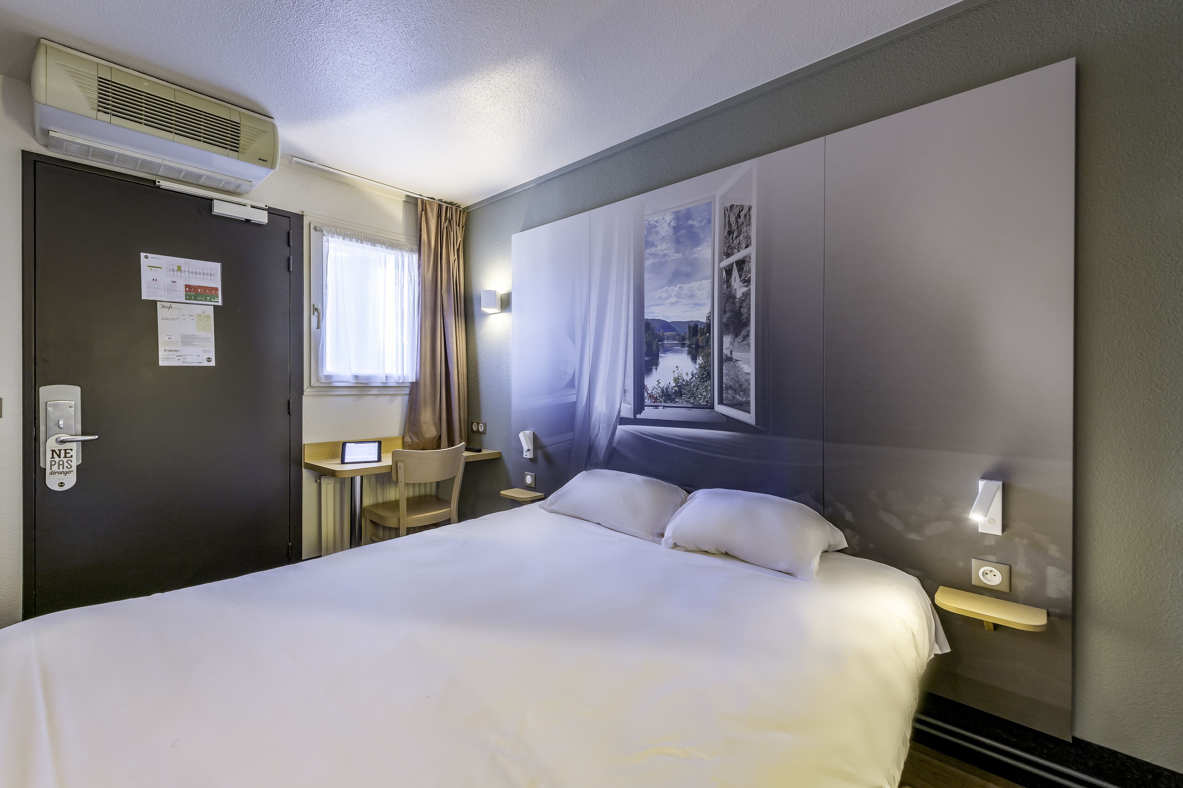 Habitación de hotel con cama, escritorio, silla, ventana con cortinas, unidad de aire acondicionado y una lámpara de pared.