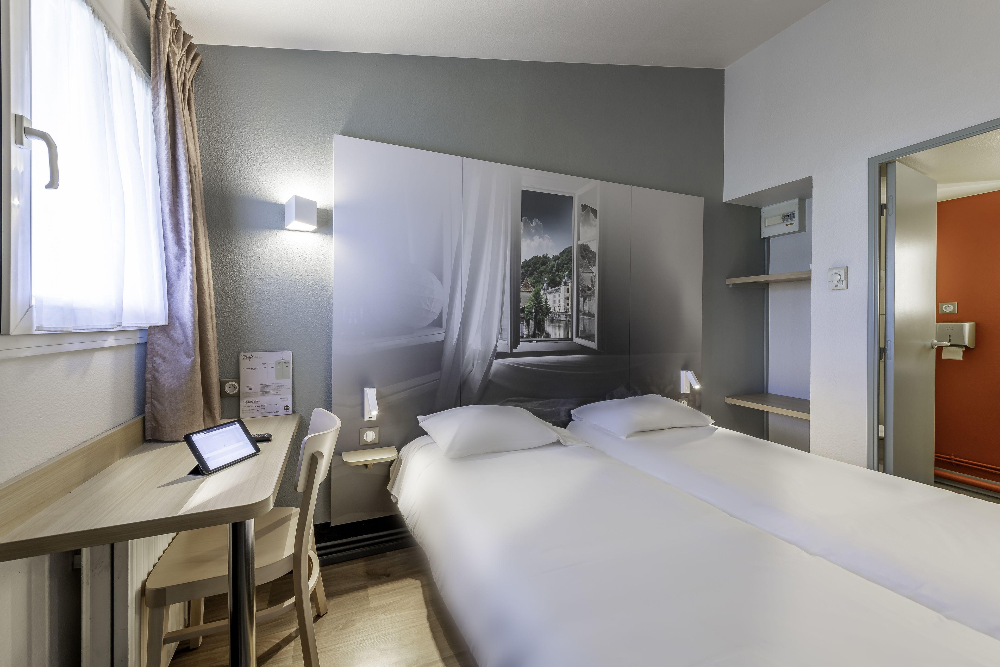 A hotel room with a bed, a desk with a tablet, a chair, a nightstand, and a wall-mounted lamp.
