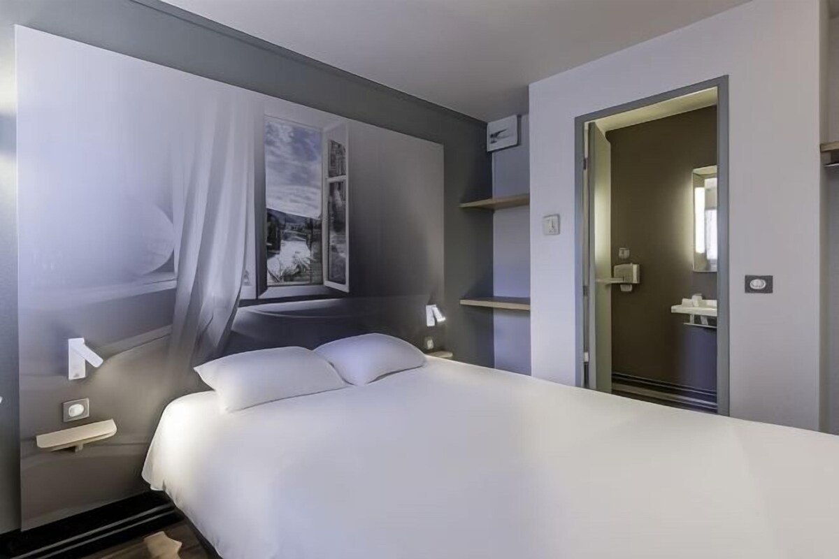 A modern hotel room with a large bed, a nightstand, a wall-mounted TV, and a door leading to a bathroom.