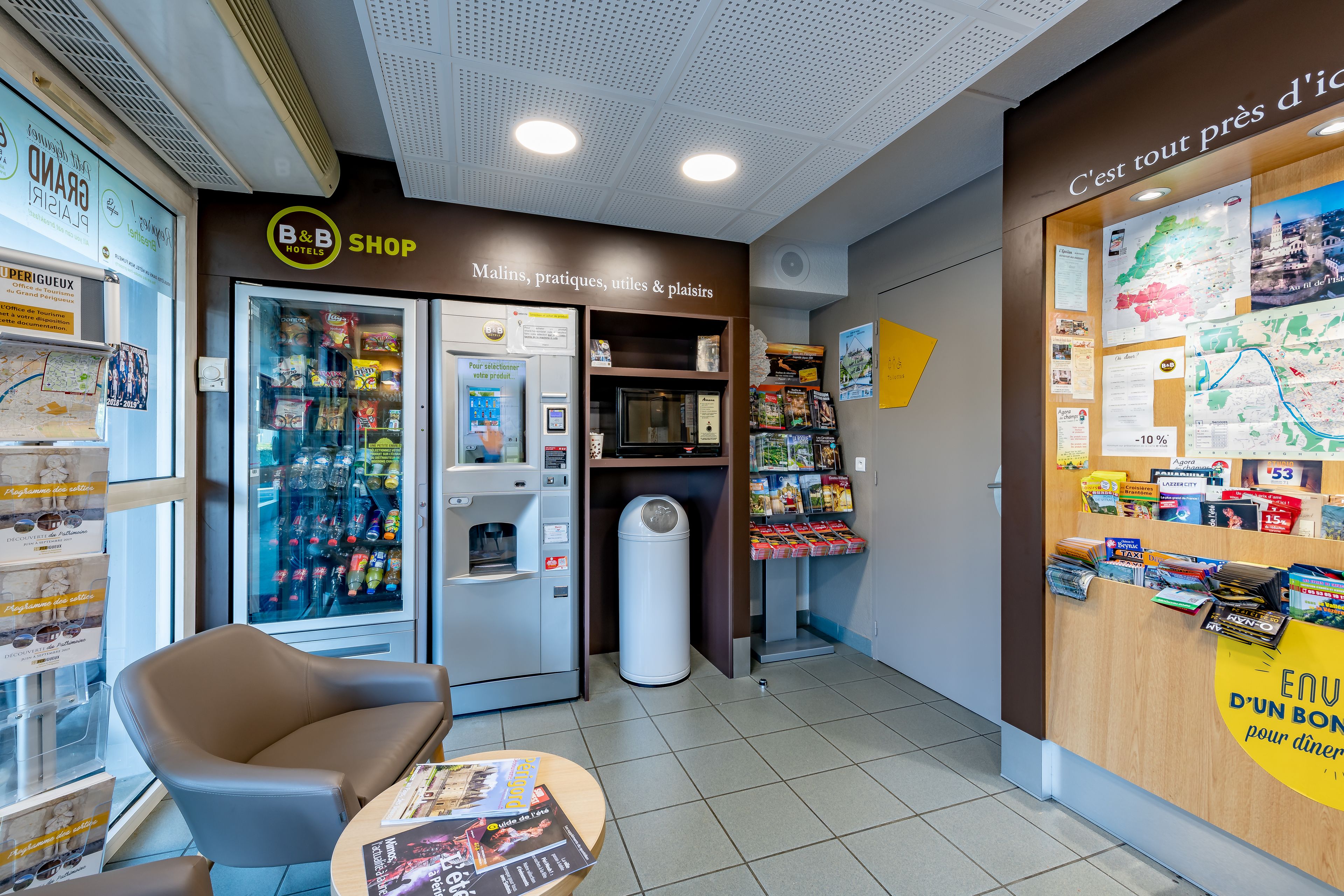 Een B&B winkel met een automaat voor zelfbediening drankjes, een prullenbak en een zithoek met een tafel.
