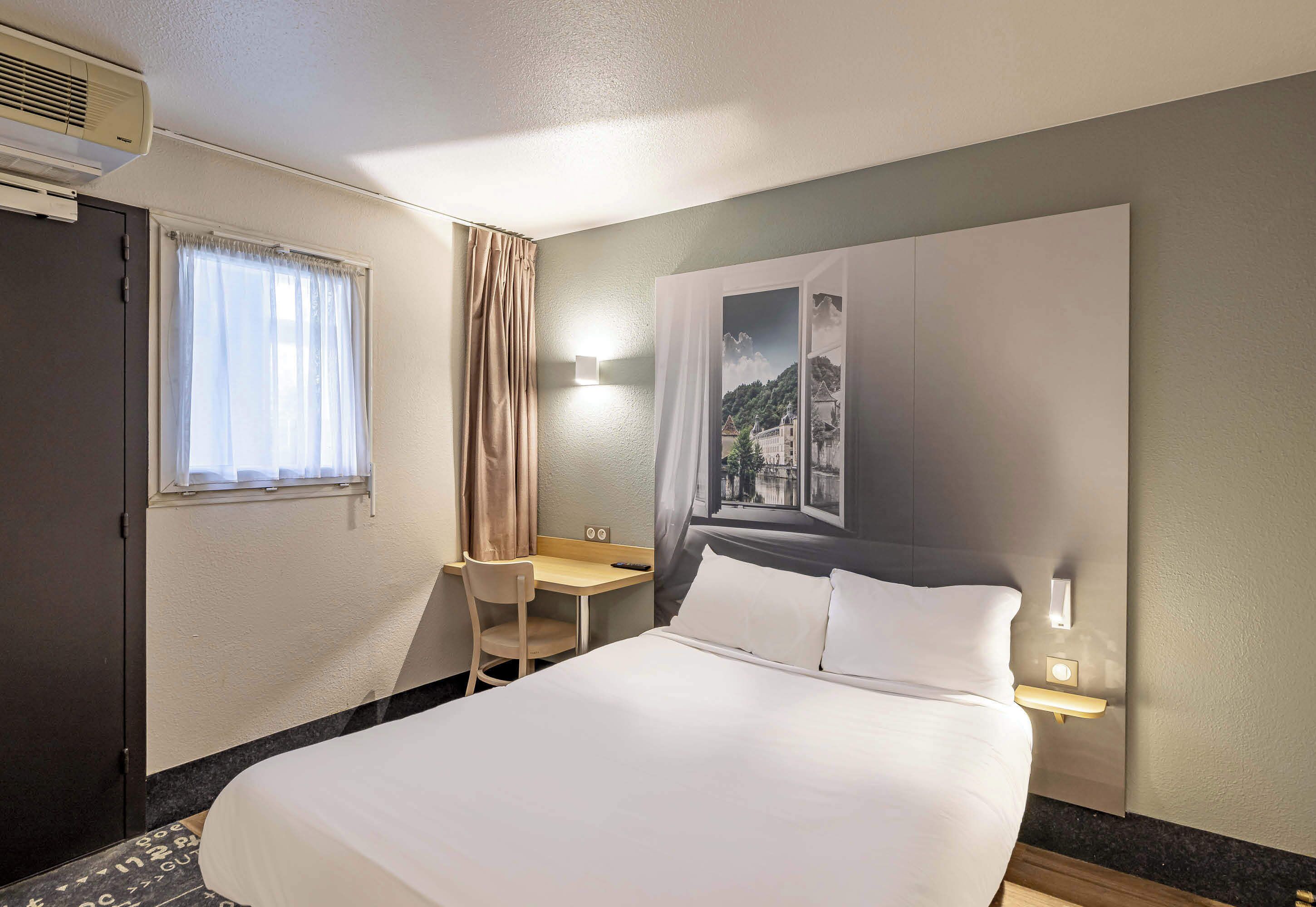 Ein Hotelzimmer mit Bett, Schreibtisch, Stuhl, einem Fenster mit Vorhängen und einer Wandleuchte.