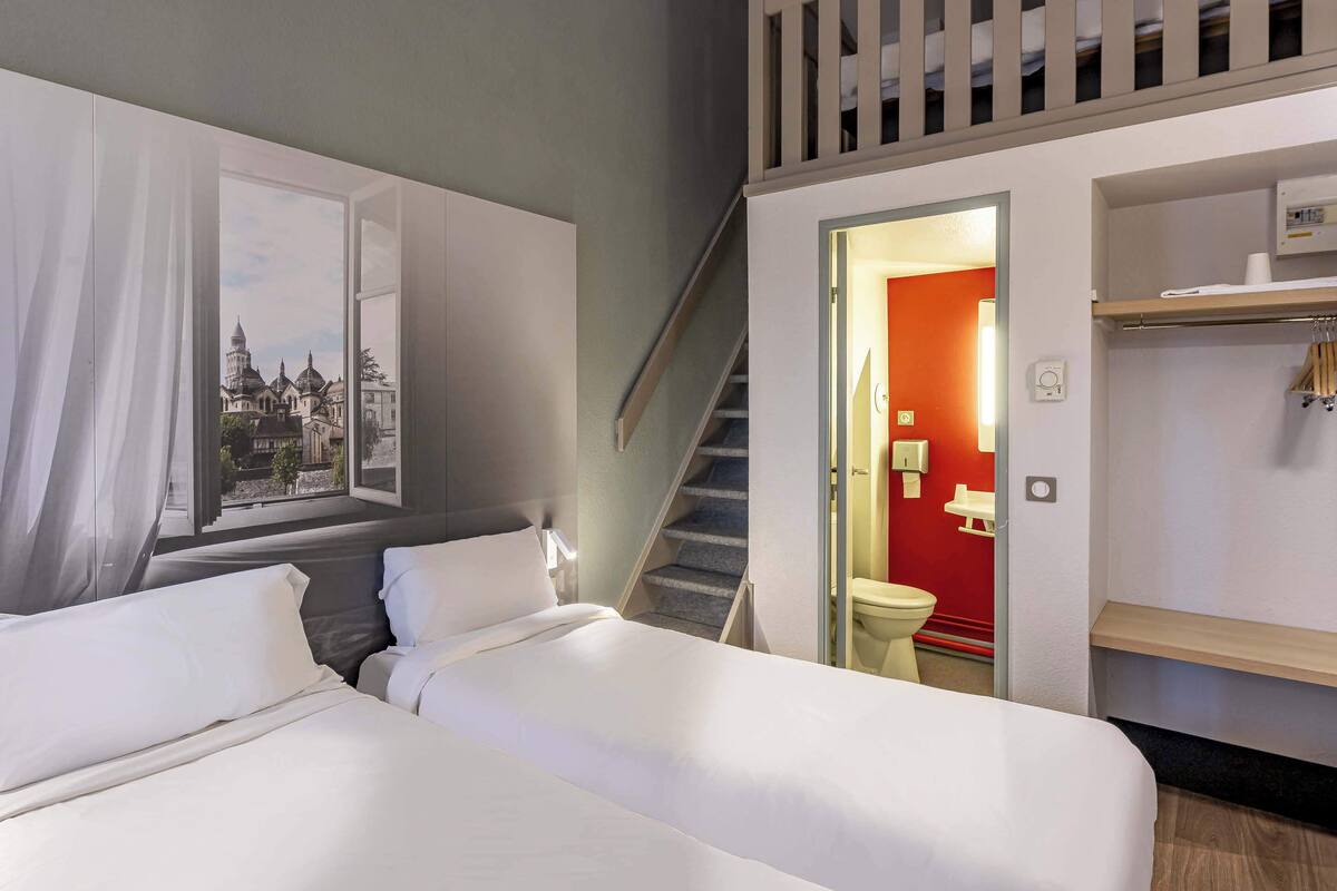 A hotel room with two beds, a staircase, and a view of a cityscape through a window.