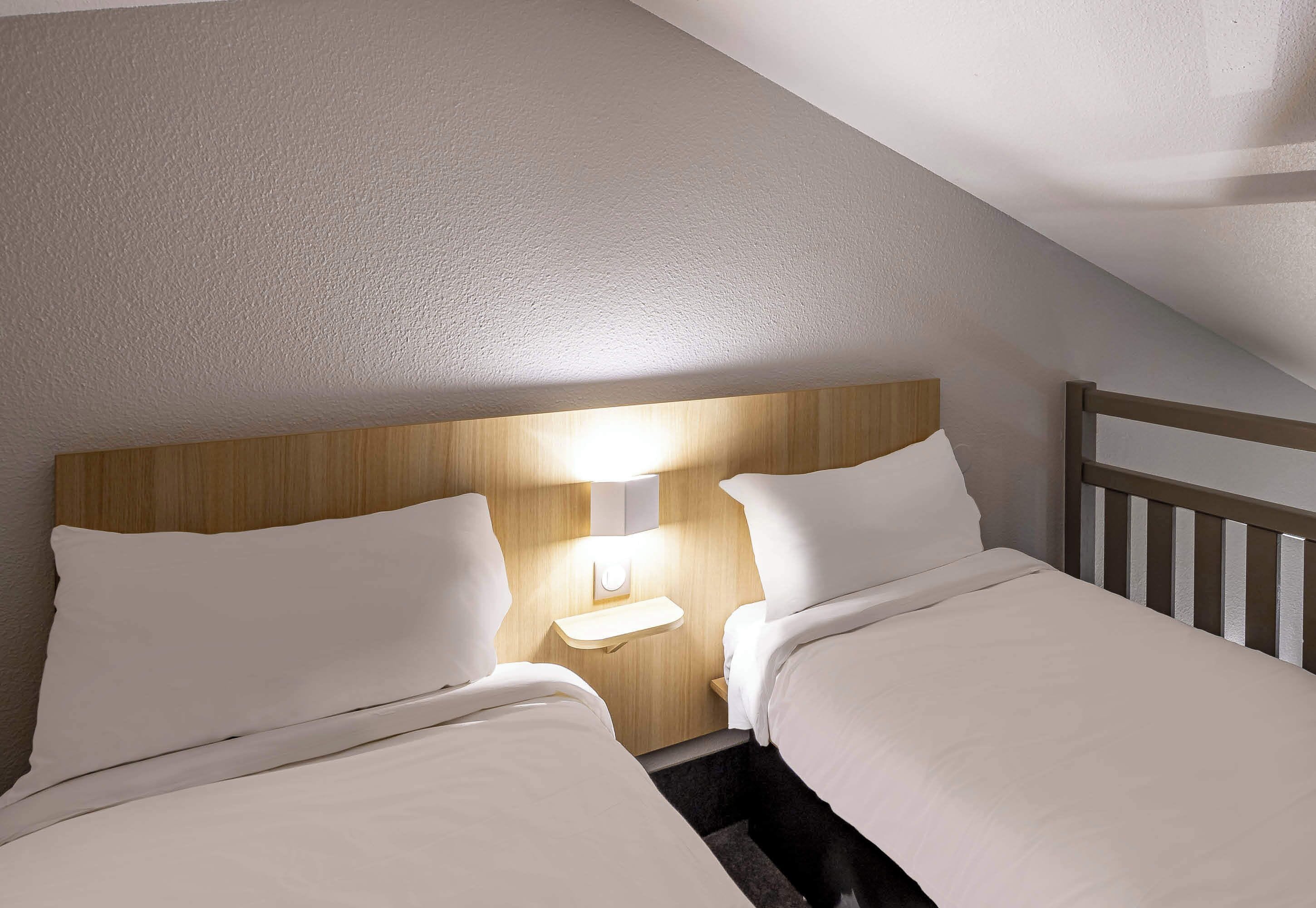 A small, cozy room with two beds, a wooden headboard, and a nightstand with a lamp.