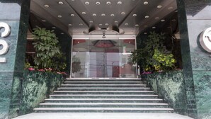 Un escalier menant à l’entrée d’un bâtiment, avec une porte en verre, le tout entouré de verdure et bordé de murs en marbre.