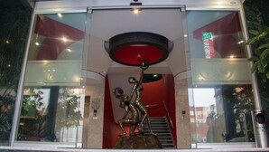 L’entrée d’un hôtel se distingue par sa façade en verre, son intérieur rouge et une statue en bronze représentant un homme et une femme.