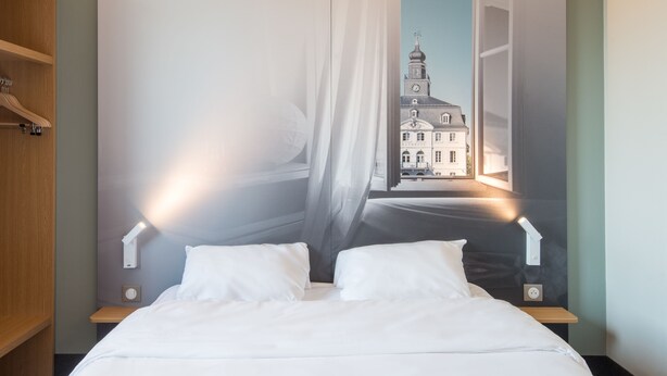 Une chambre à coucher comprenant un lit, deux tables de chevet, une commode, une lampe fixée au mur et une grande fenêtre donnant sur un immeuble.