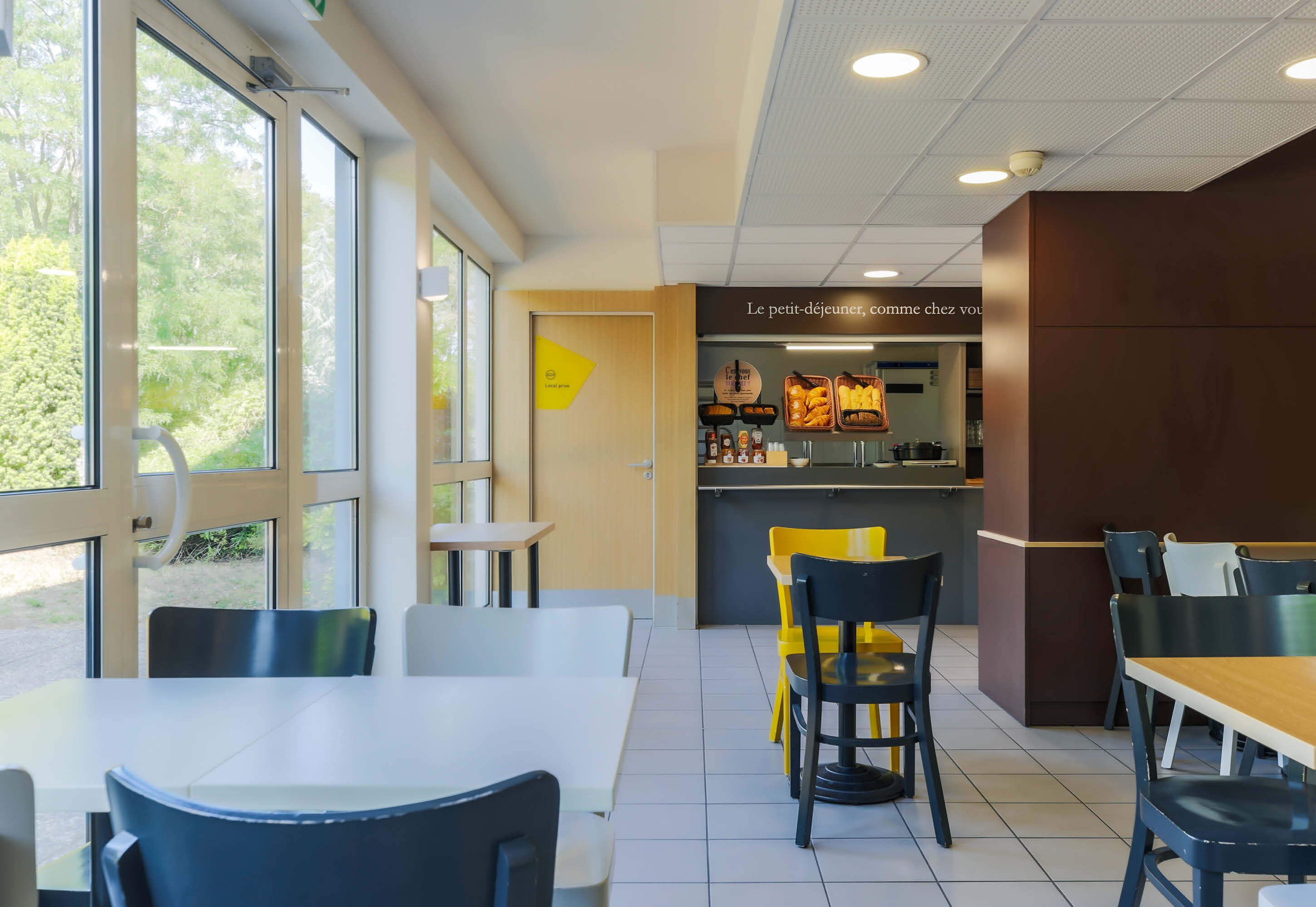 A modern restaurant interior with a dining area, a counter with food display, and large windows offering a view of greenery.