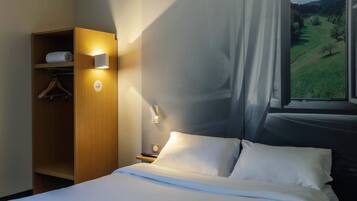 A hotel room with a bed, a wooden wardrobe, a nightstand, and a window with a view of a green landscape.