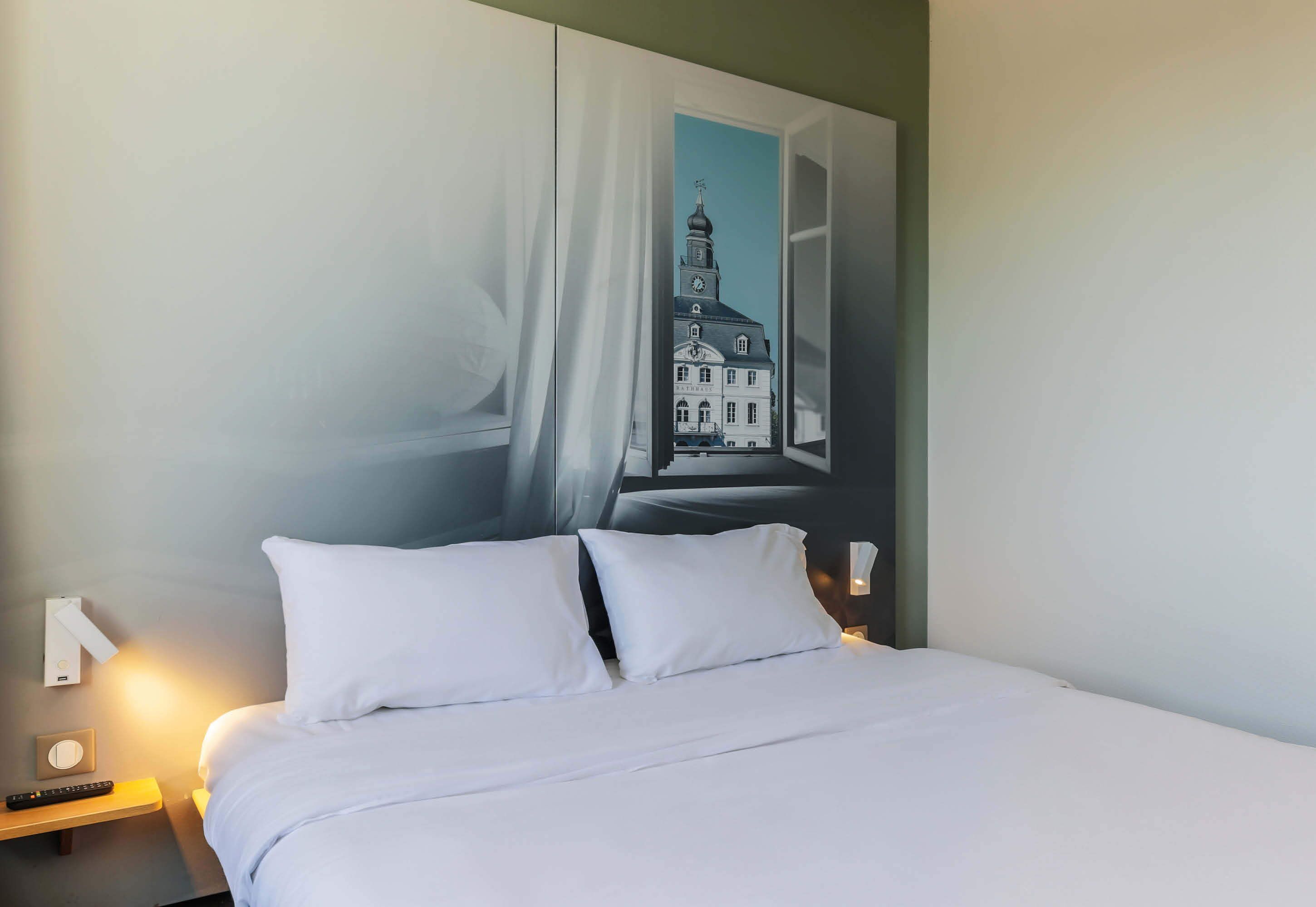 Habitación de hotel con cama, ropa de cama blanca, mesita de noche con lámpara y un cuadro colgado en la pared de un edificio.