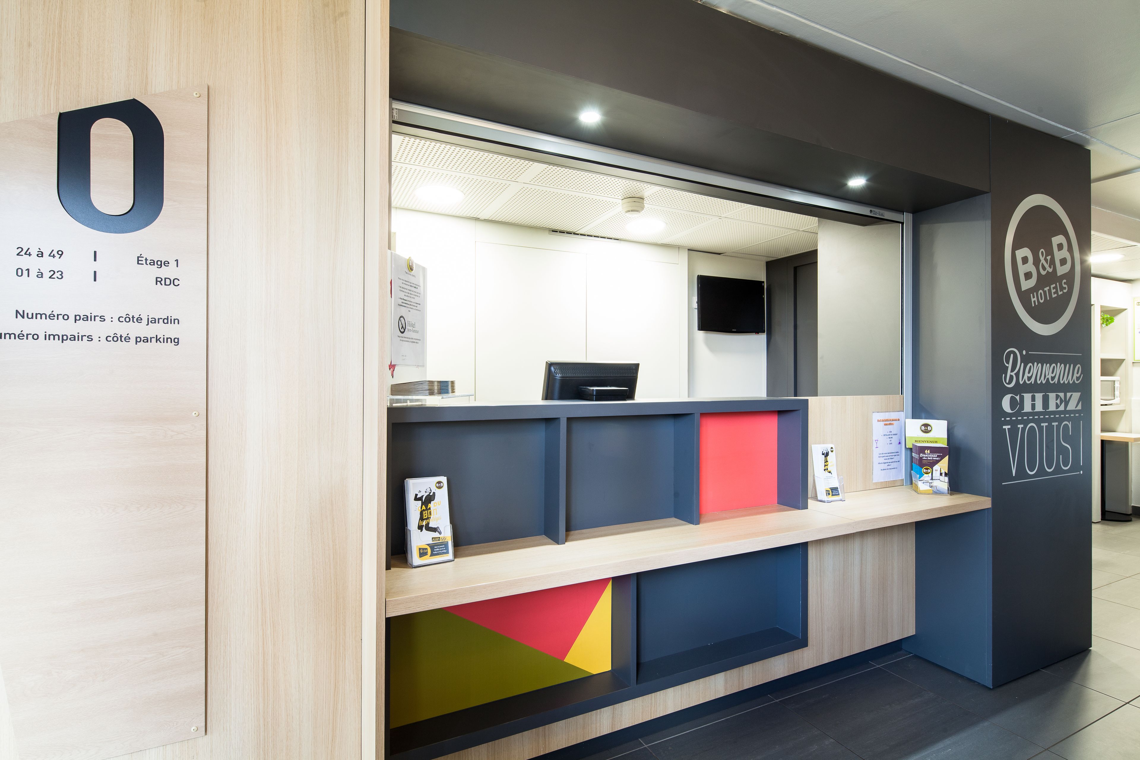Un comptoir d’accueil avec un panneau indiquant les numéros des chambres et une enseigne d’accueil de la chaîne B&B Hotels.