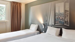 A hotel room with two single beds, a large window with a view of a cityscape, and a wall-mounted lamp. - B&B HOTEL Corbeil-Essonnes (Corbeil-Essonnes)