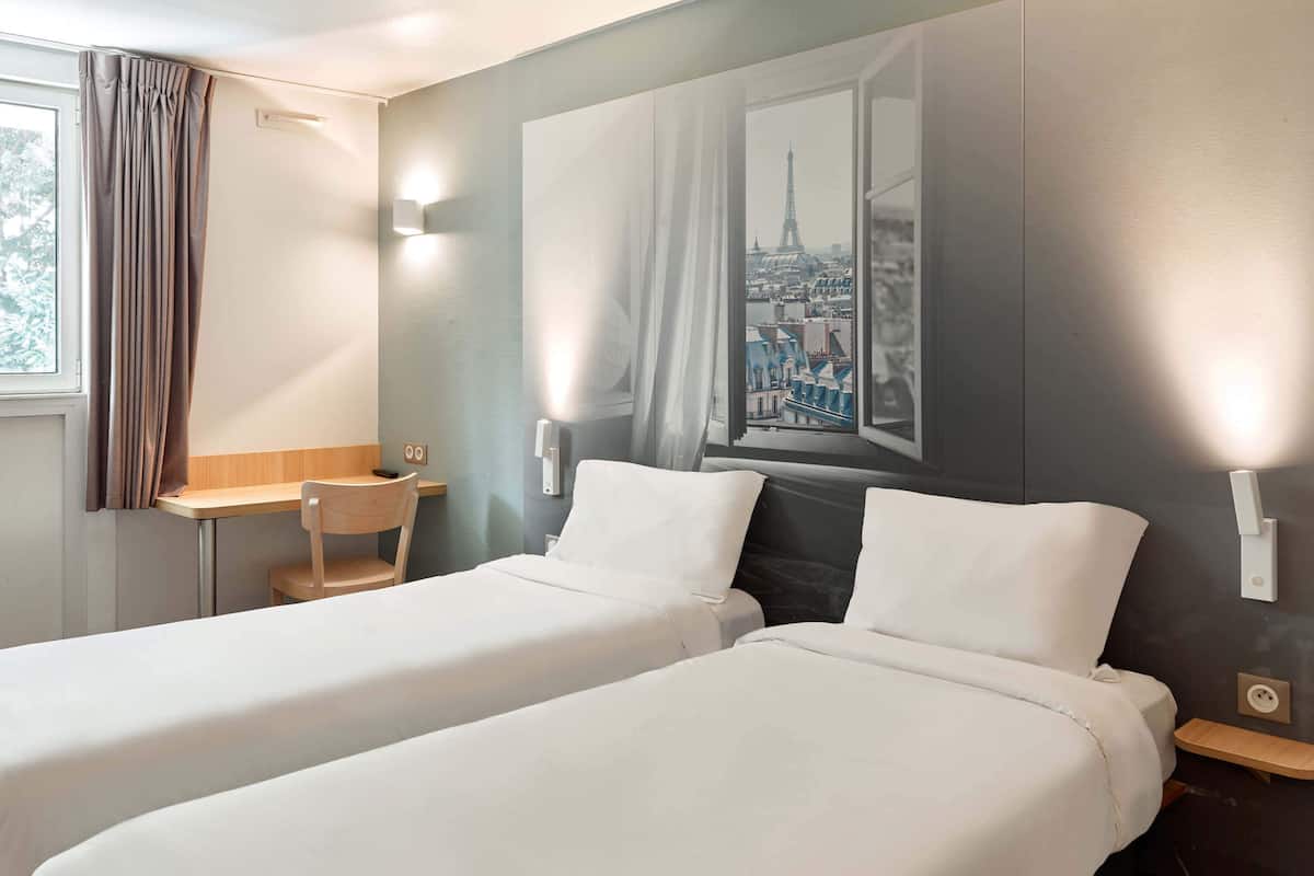 Habitación de hotel con dos camas individuales, un escritorio, una silla y un ventanal con vistas a la ciudad.