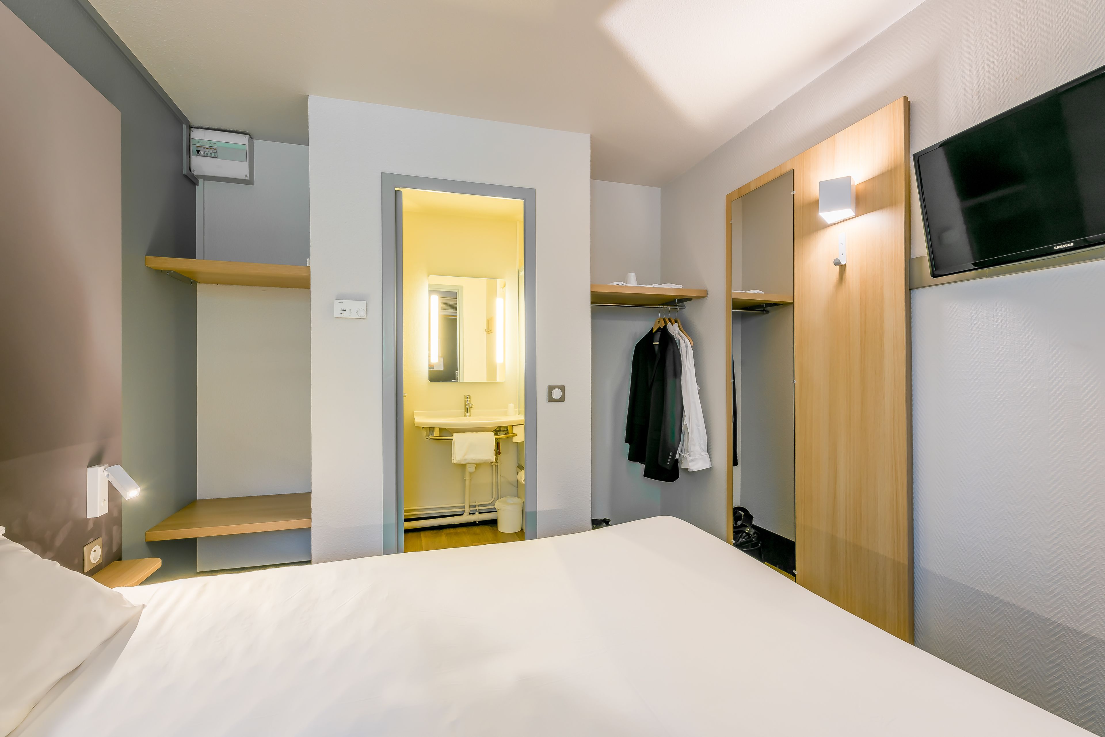 Une chambre d’hôtel avec un lit, une télévision, une armoire, un miroir et une salle de bain visible à travers une porte ouverte.
