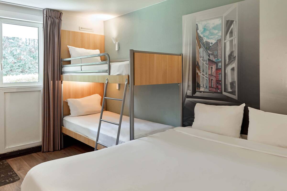 Una habitación de hotel con literas, una cama individual, una escalera, una ventana con cortinas y un cuadro en la pared.