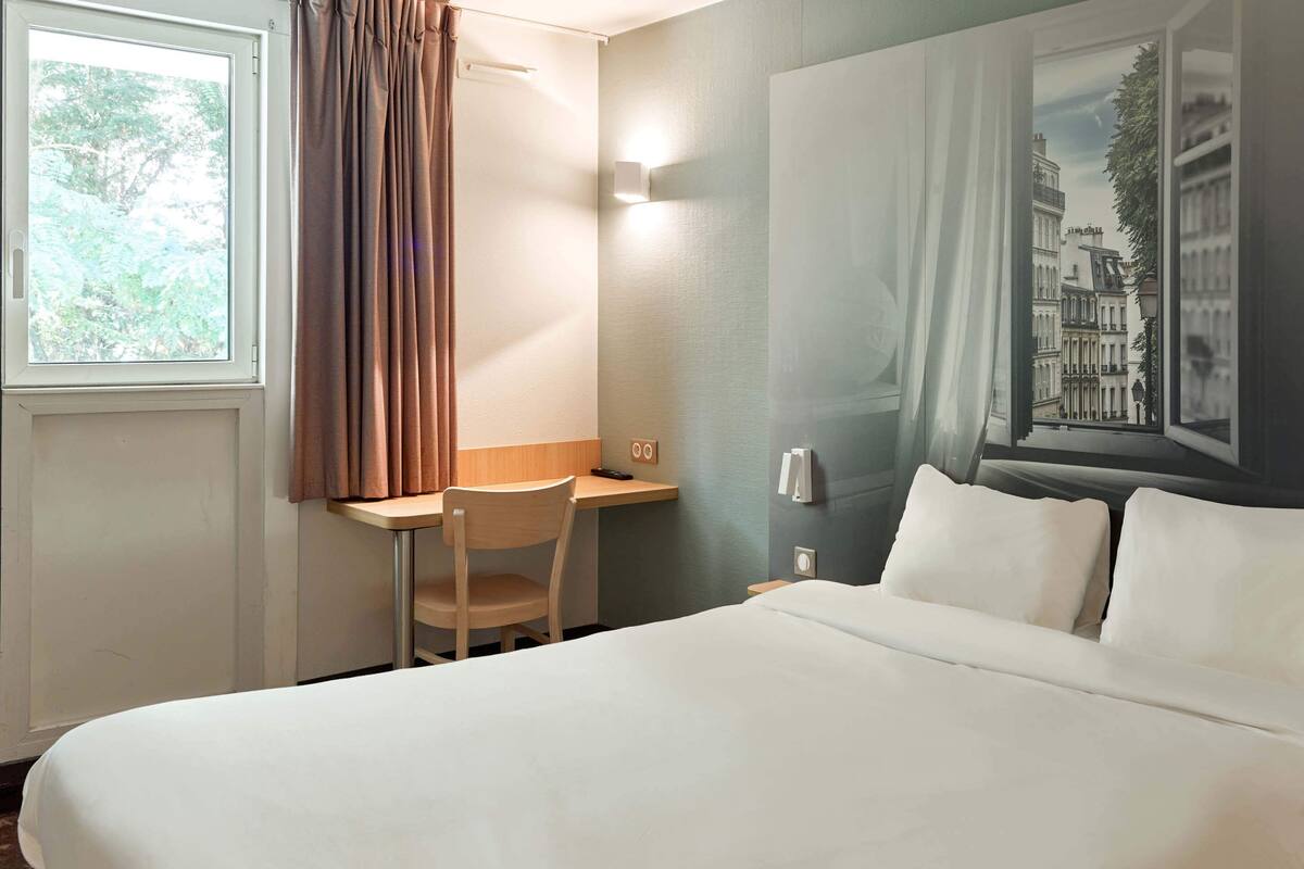 Habitación de hotel con cama, escritorio, silla, ventana con cortinas y una lámpara fijada a la pared.