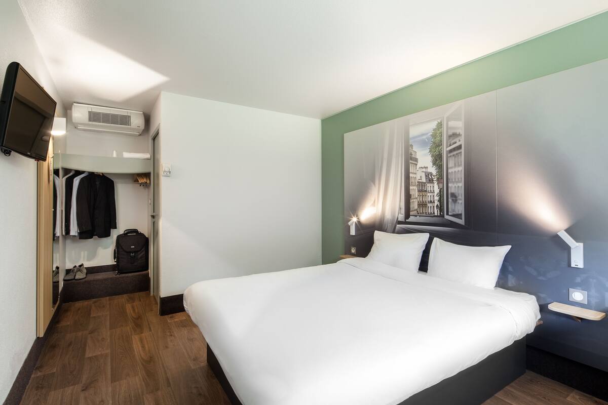 Habitación de hotel con una cama grande, un televisor de pantalla plana montado en la pared, un armario y una pequeña zona de estar con una mesa.