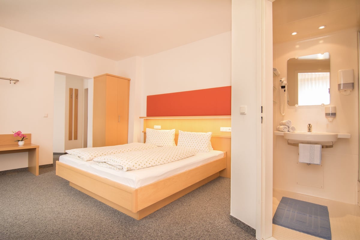 Una habitación de hotel moderna con una cama de madera, un pequeño escritorio, un baño con lavamanos y espejo, y una zona de ducha.