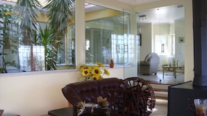 Una sala de estar con un sofá de cuero, una mesa de centro de madera y un jarrón con girasoles. Amplias ventanas ofrecen vistas a palmeras y un espejo refleja la habitación.