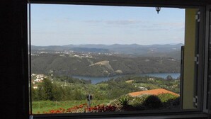 A room with a large window offering a view of a lake, hills, and a red-roofed building.