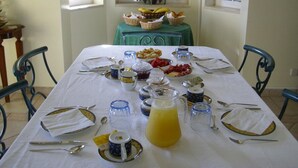 Mesa de desayuno servida con pan, fruta y bebidas, en una habitación con ventanas y un jarrón de flores.