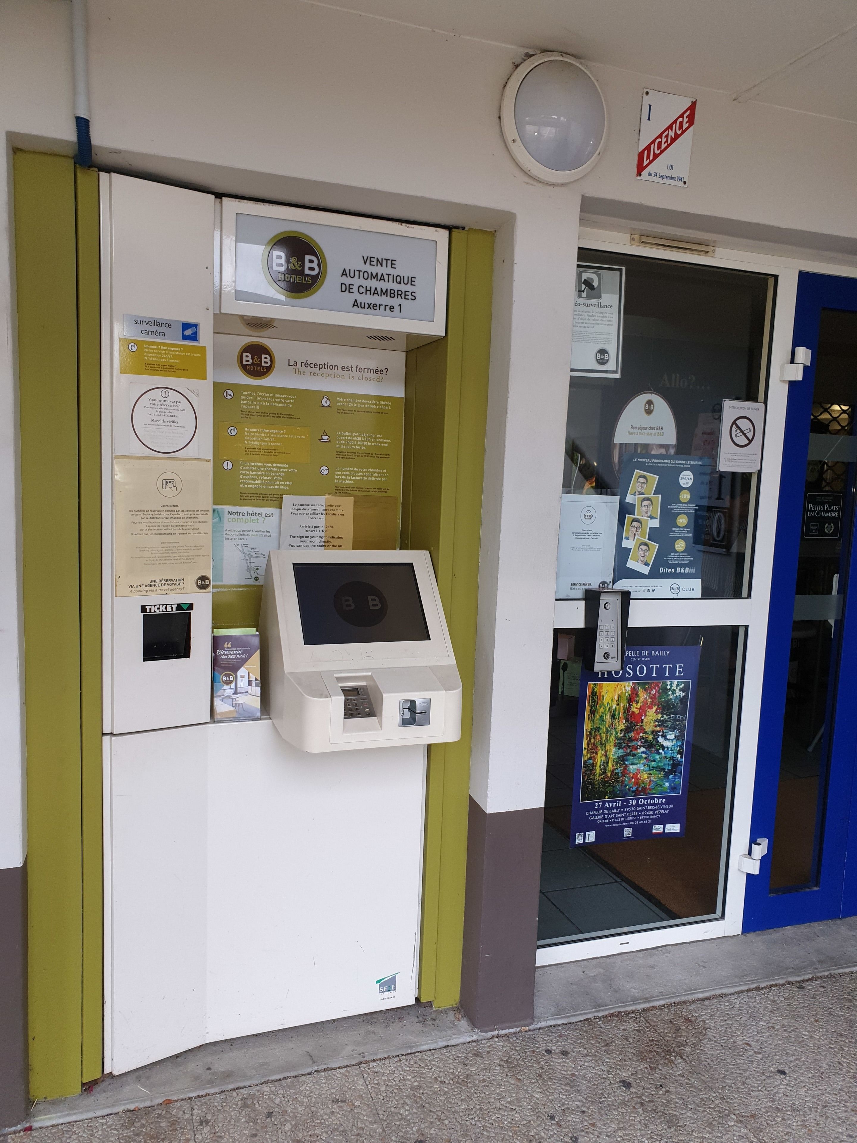Zona de recepción de un hotel con una máquina de venta de billetes, carteles informativos y una puerta de recepción cerrada.