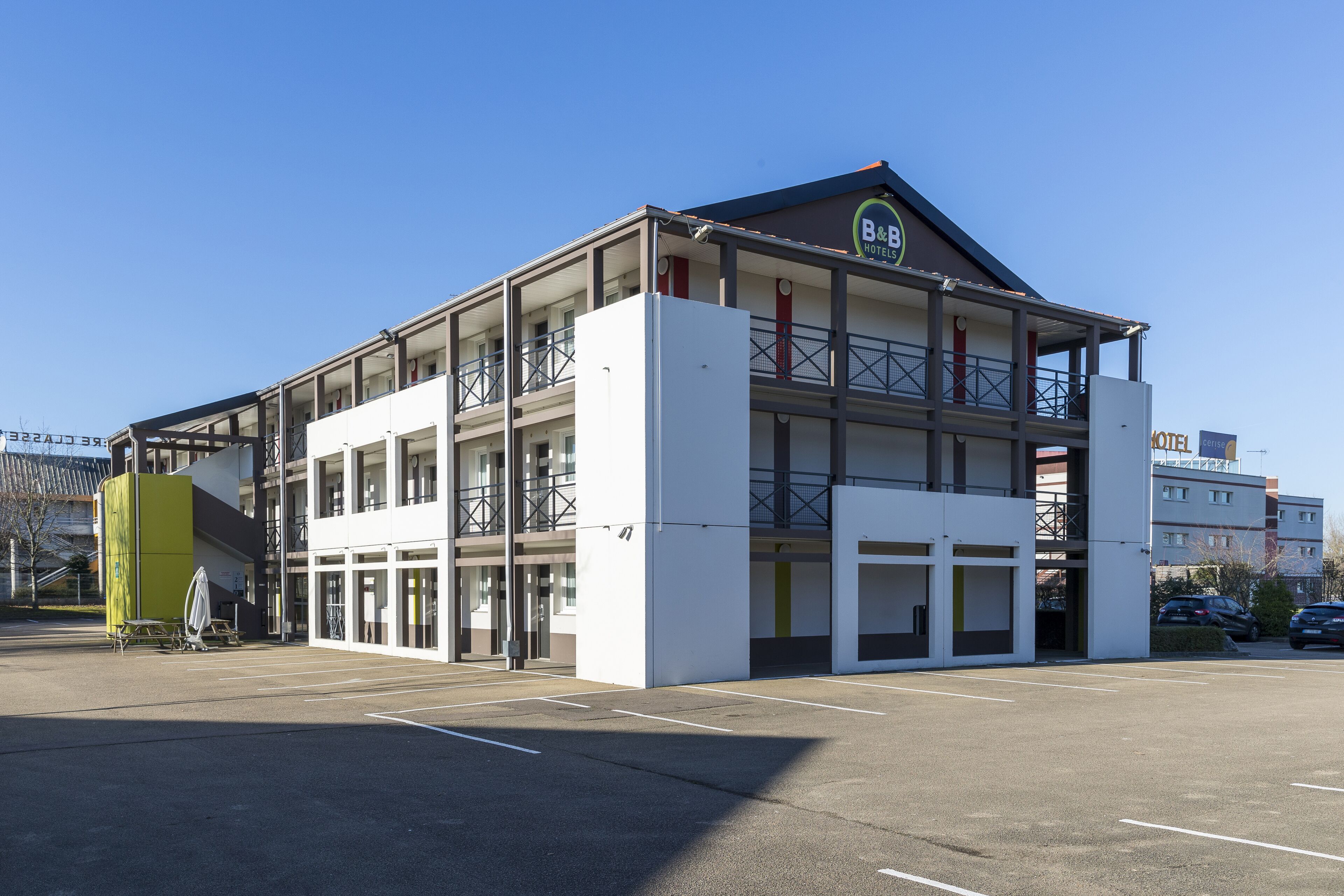 A two-story building with a flat roof and a balcony on the second floor. There are multiple windows on each floor, and the building has a sign that reads "B&B Hotels".