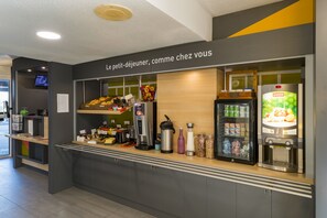 Un espace de petit-déjeuner en libre-service comprenant une machine à café, un réfrigérateur rempli de boissons, ainsi qu’un présentoir de fruits frais et de viennoiseries.