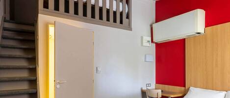 A hotel room with a bed, a desk, a chair, a staircase, and a red accent wall.