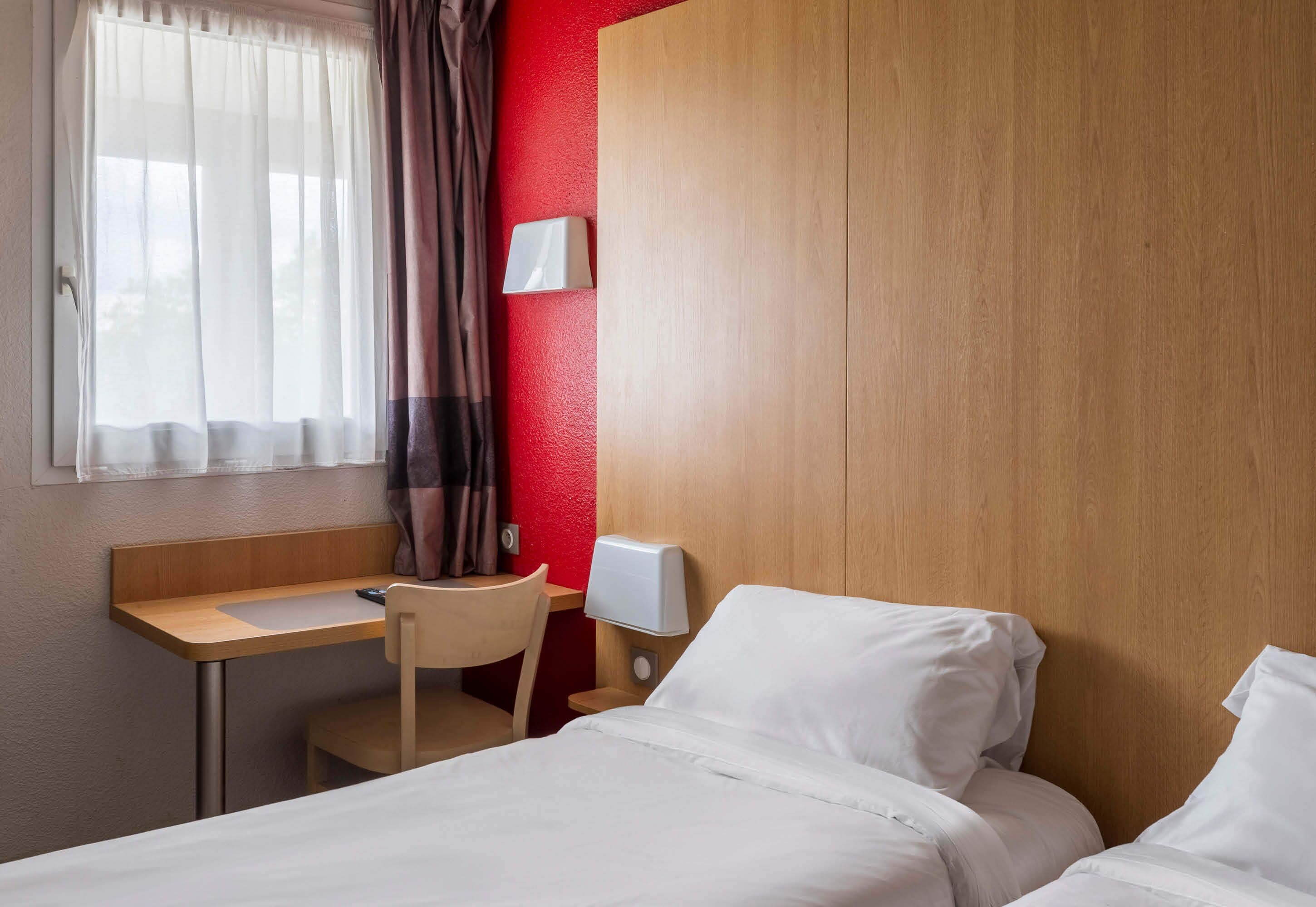A hotel room with a wooden desk, a chair, a bed with white linens, a bedside table with a lamp, and a window with sheer curtains.
