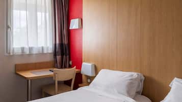 A hotel room with a wooden desk, a chair, a bed with white linens, a bedside table with a lamp, and a window with sheer curtains.