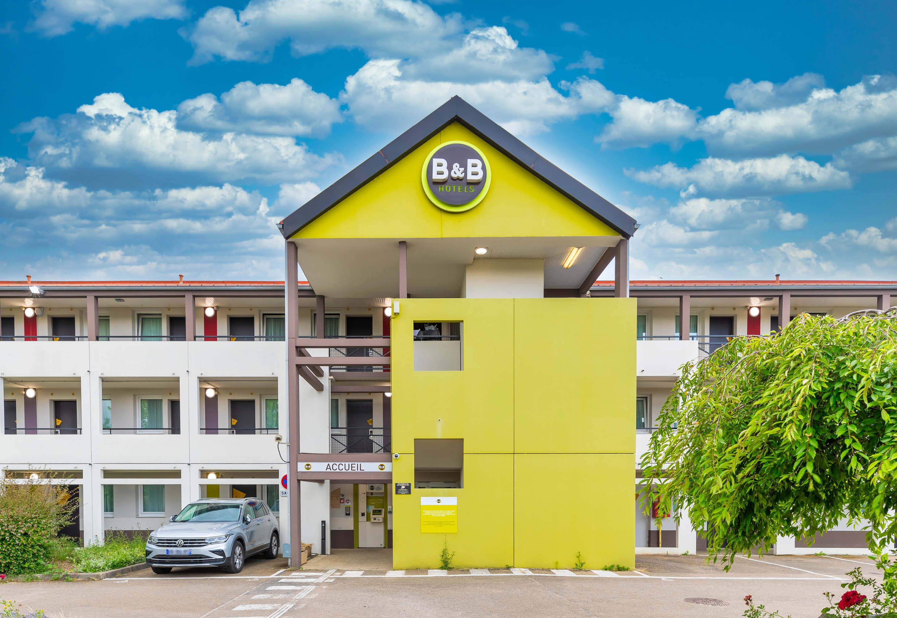 A B&B hotel with a yellow facade, a parking area, and a sign indicating an entrance.