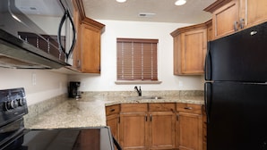 A kitchen with wooden cabinets, a black refrigerator, a stove, and a window with blinds. - Duchesne Suites (Duchesne)