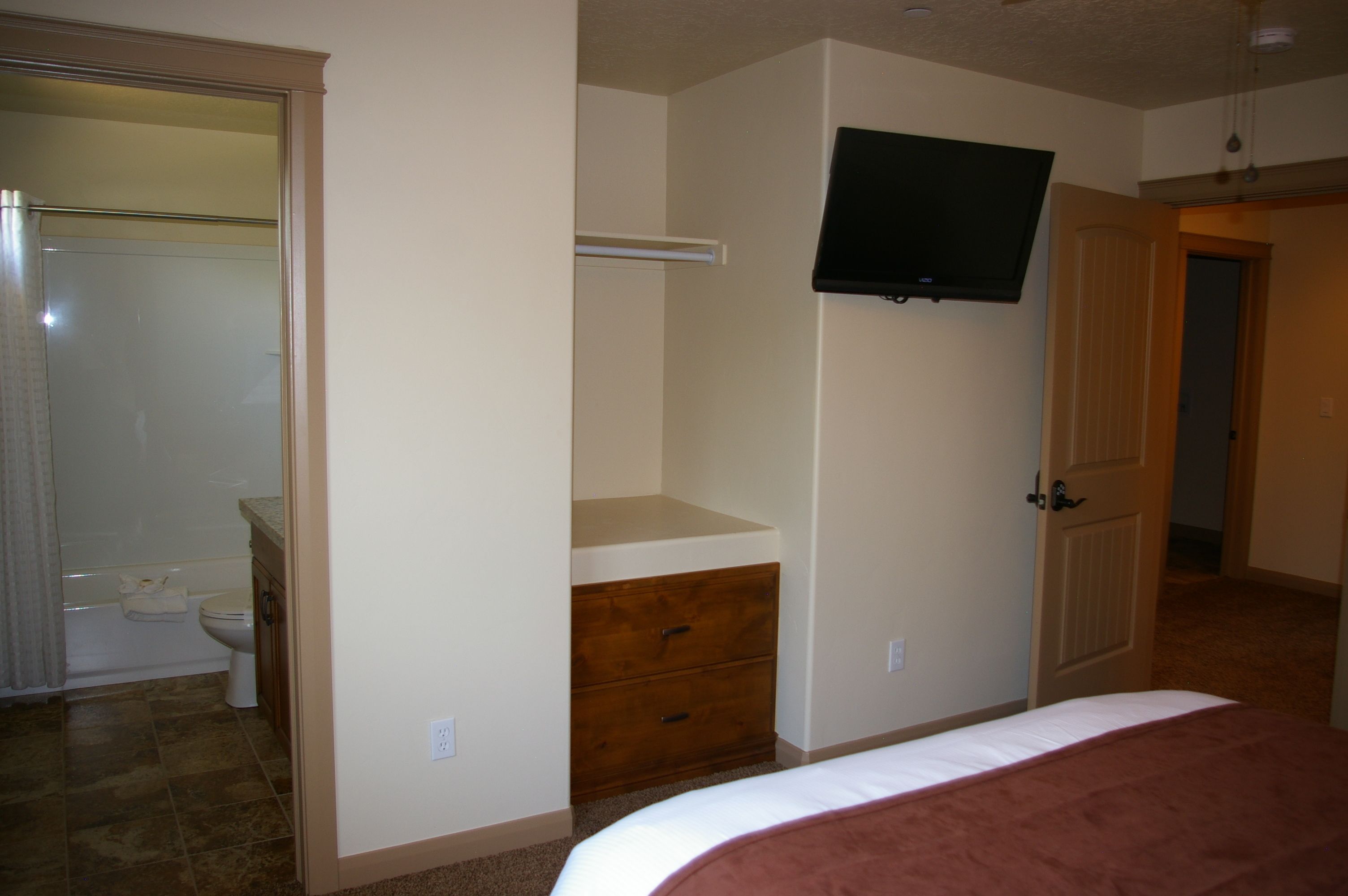 Un dormitorio con cama, cómoda, televisor montado en la pared y una puerta que da a otra habitación.