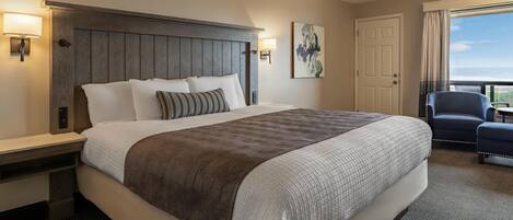 Habitación de hotel con una cama grande, cabecera de madera, mesita de noche, escritorio, silla y vista al océano.
