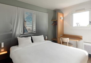 A hotel room with a bed, a desk, a chair, a window with a view, and a coat rack. - B&B HOTEL Montlhéry (Linas)