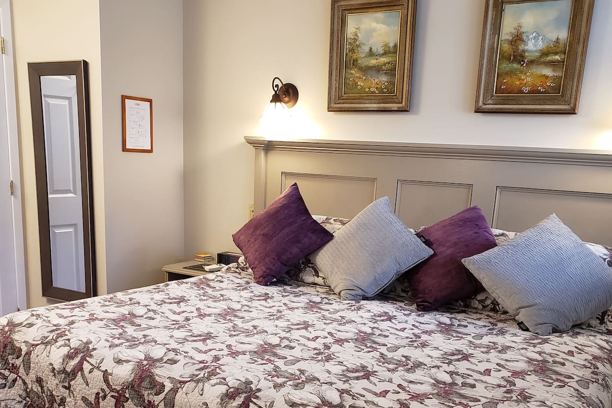 Une chambre avec un grand lit, une table de chevet, une lampe, deux tableaux encadrés au mur et un banc placé devant le lit.