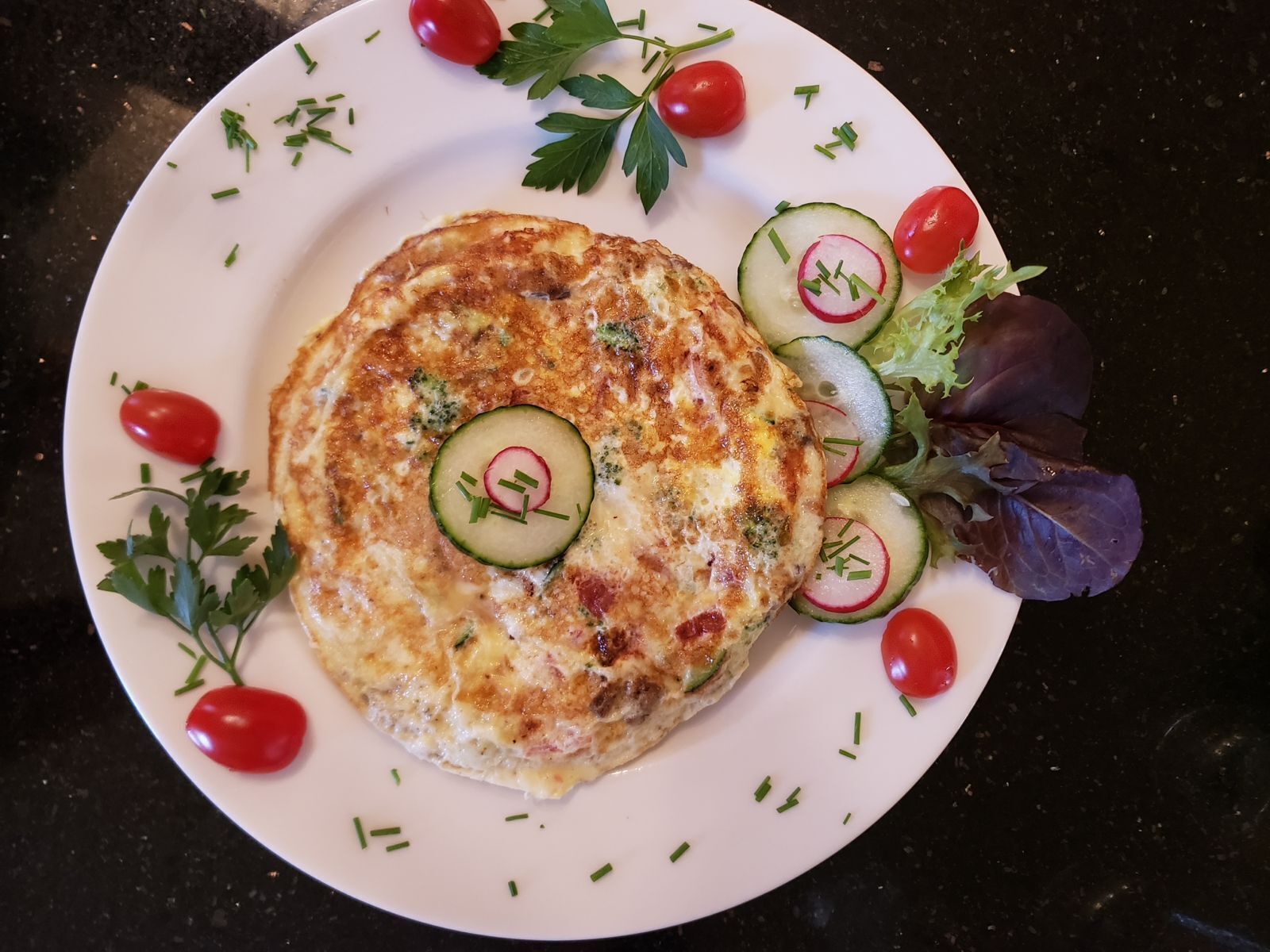 A plate with a vegetable omelette, garnished with cherry tomatoes, cucumber slices, and parsley.