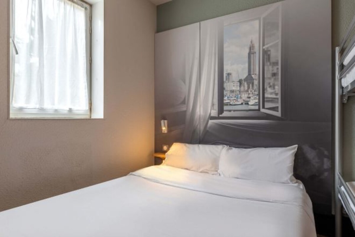 Una habitación de hotel con una cama, una ventana con cortinas, un mural en la pared y una mesita de noche con una lámpara.