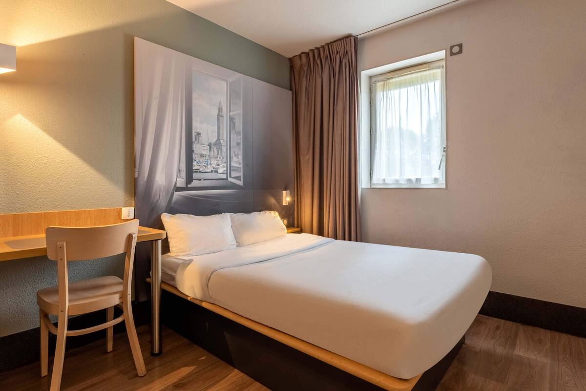 A hotel room with a bed, a desk, a chair, a window with curtains, and a wall with a cityscape painting.