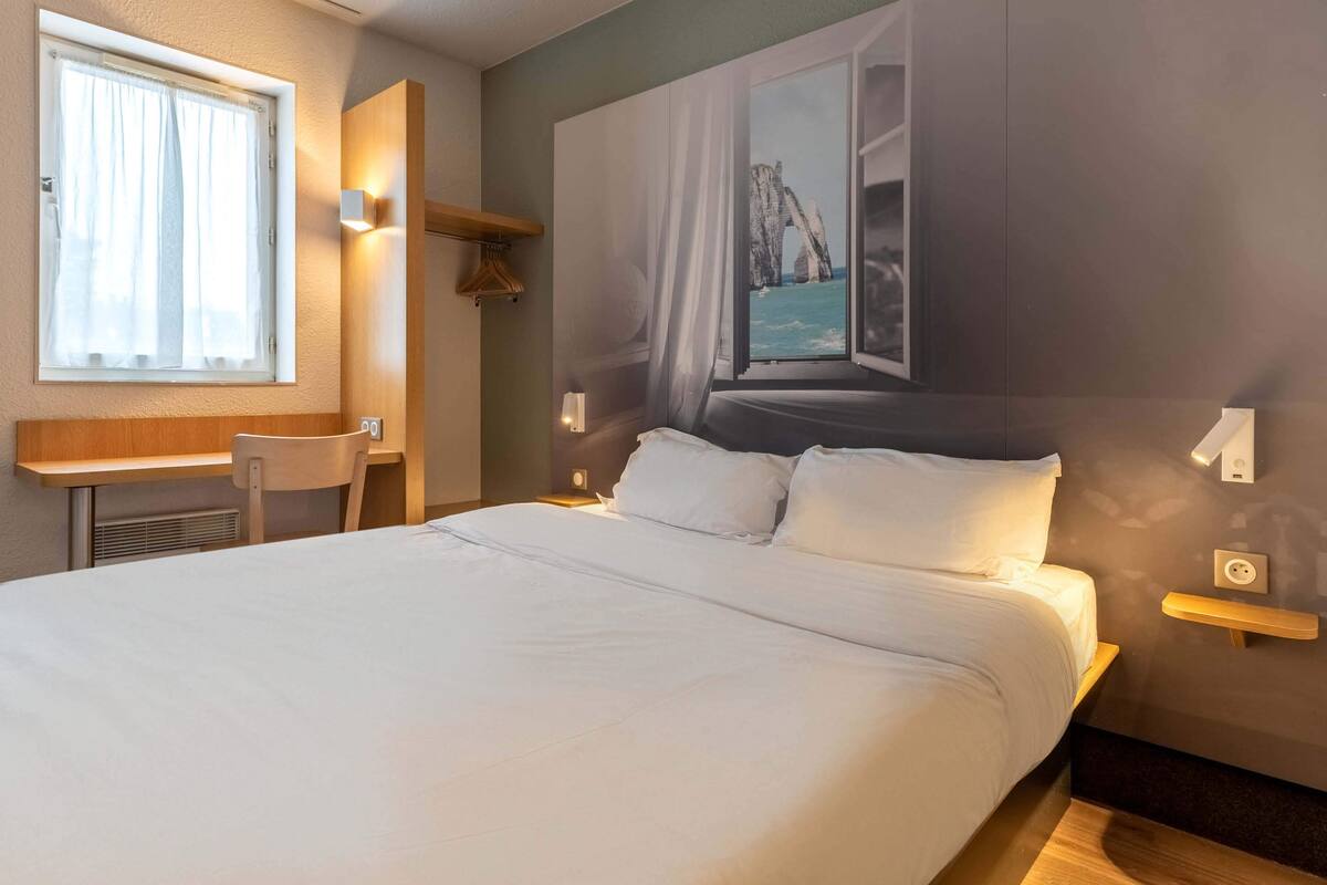 A hotel room with a large bed, a desk, and a chair. There is a window with curtains, a wall-mounted shelf, and a painting of a coastal scene.