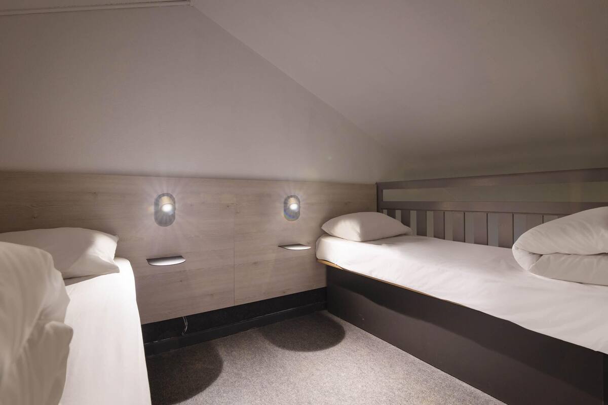 A compact, well-lit room with two beds, a wooden headboard, and built-in wall lighting.