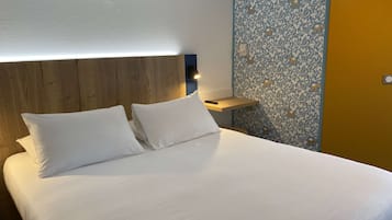 Un lit bien fait, avec du linge de lit blanc et deux oreillers, une tête de lit en bois, une table de chevet avec une lampe et un mur orné d’un motif floral.