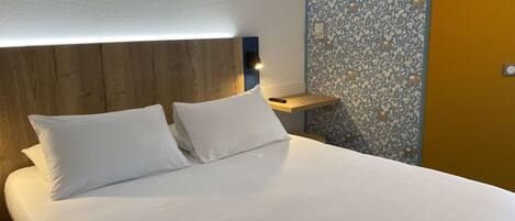 A neatly made bed with white linens and two pillows, a wooden headboard, a bedside table with a lamp, and a wall with a floral pattern.