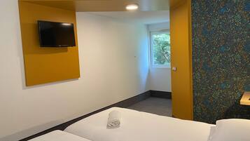 A hotel room with a bed, a television mounted on a yellow wall, and a window with a view of trees.